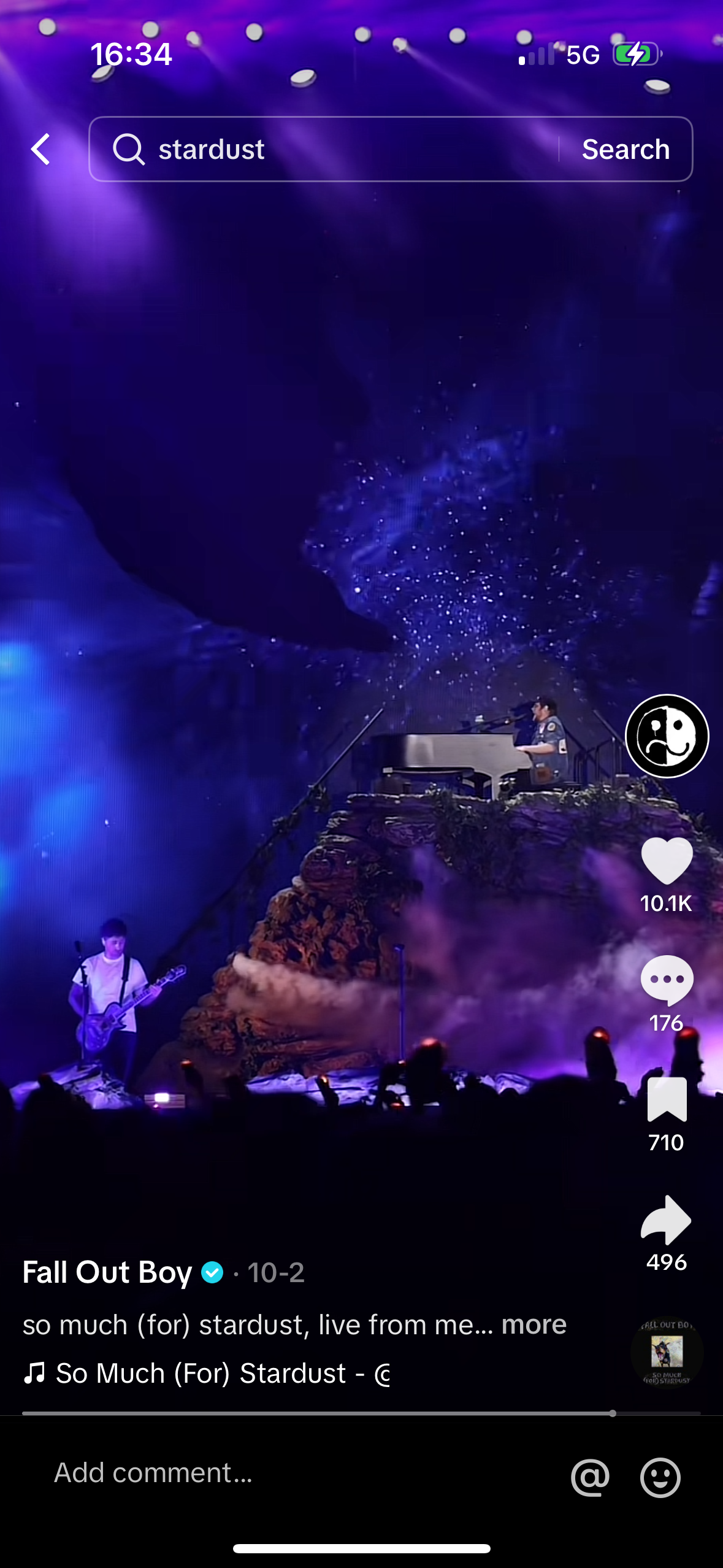 a live stream of the band fall out boy's concert performing the song "so much (for) stardust". the stage is set with a piano and drums at the front and a singer standing behind it with a microphone. the singer is playing a guitar and the rest of the band is visible on the screen, but blurred. there are people in the audience who are all blurred and obscured by the stage lights. there are a few social media icons displayed on the screen including a heart with a number under it, a chat bubble with a number under it and a notification button that says "710". there are also playback controls.