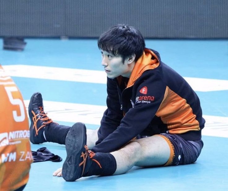 a young male volleyball player in an orange and black uniform sits on the court floor, stretching his legs. he is wearing black volleyball shoes. the image is taken from a closeup perspective.