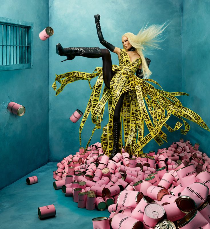 the image is a highfashion, conceptual photograph featuring a blonde model in a striking pose. she's wearing a dress constructed entirely of yellow caution tape, her pose is a dynamic high leg kick. the model is set against a teal backdrop reminiscent of a prison cell, surrounded by a large pile of pink cans labeled with words like "processed" and "chemicals." some cans are also flying in the air, contributing to the dynamic movement in the image. the overall aesthetic is avantgarde and surreal, possibly commenting on consumerism, environmental issues or societal pressures. the contrast between the bright yellow tape dress and the pink cans against the muted teal background is visually striking.
