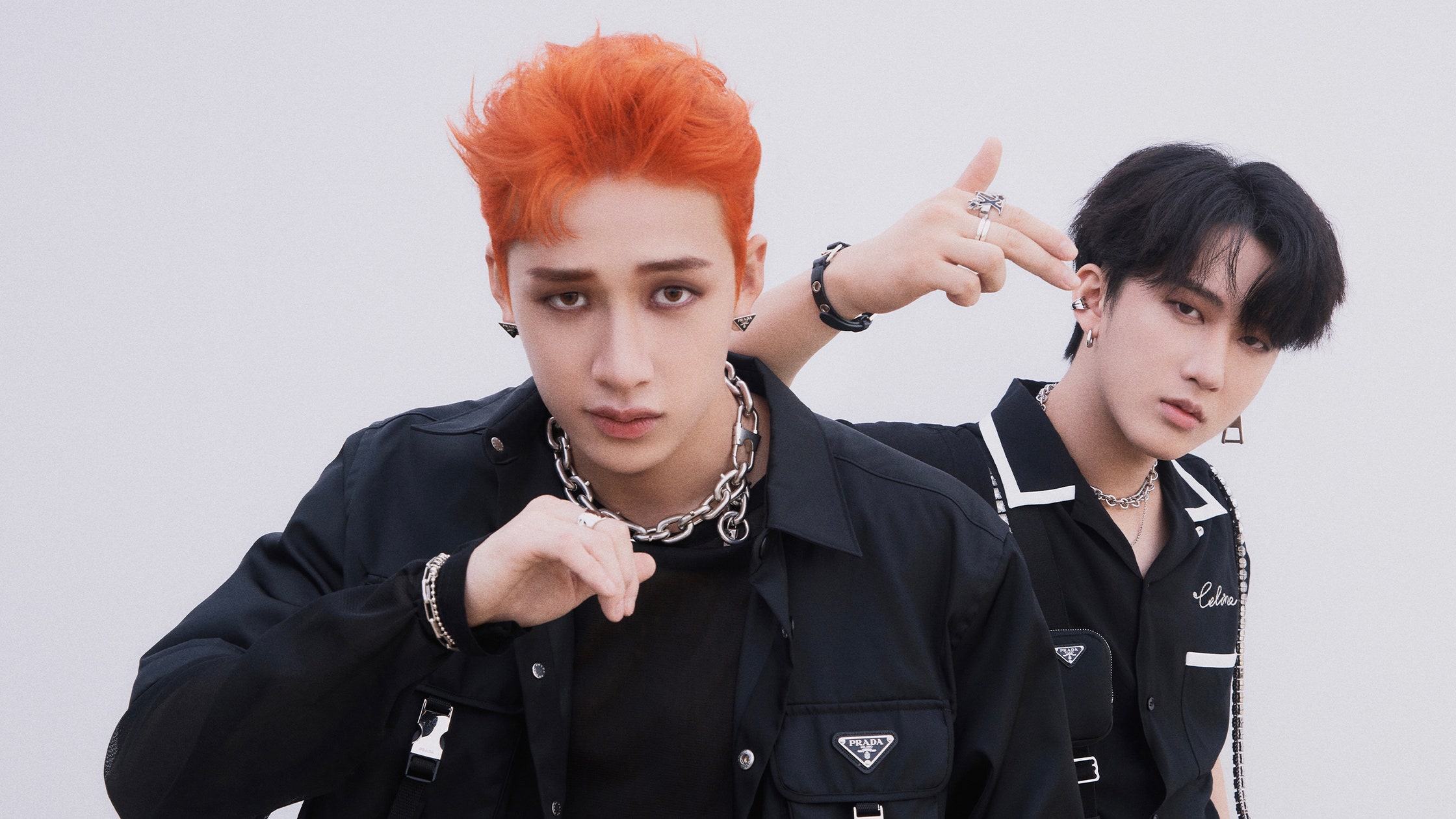 the image shows two members of the south korean boy band stray kids, bang chan (on the right with dark hair) and lee felix (on the left with bright orange hair). they are posing for a closeup shot, both wearing stylish allblack outfits accented with various silver chains, rings, and earrings.  felix's hair is a striking orange.  bang chan is playfully pointing a finger at felix's head. the overall aesthetic is edgy and trendy. the background is a simple, light gray, keeping the focus on the idols.