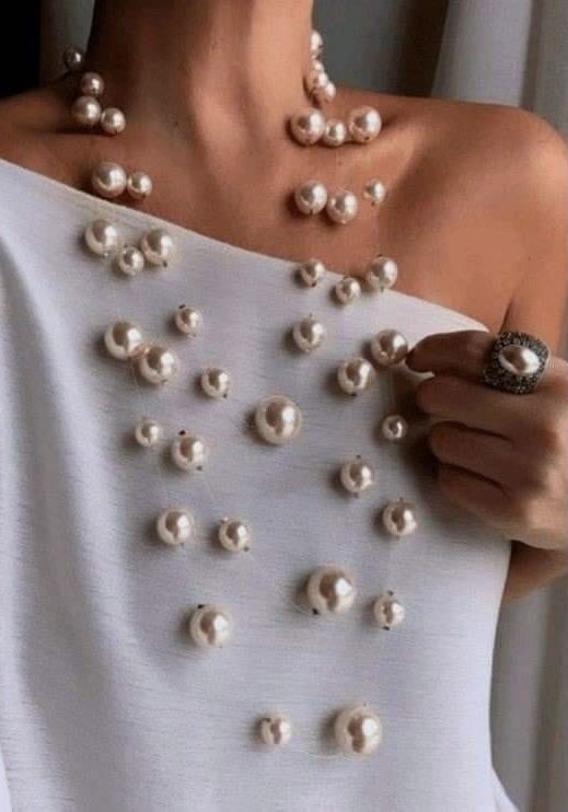 a woman is wearing a white offtheshoulder shirt adorned with a cascading arrangement of large pearls. she is also wearing a dark ring on her index finger. the pearls create a visually striking and elegant statement piece. the lighting is soft and indoor, highlighting the luster of the pearls and the smooth texture of the shirt. the closeup perspective emphasizes the intricate details of the pearl arrangement and the woman's stylish choice of jewelry.
