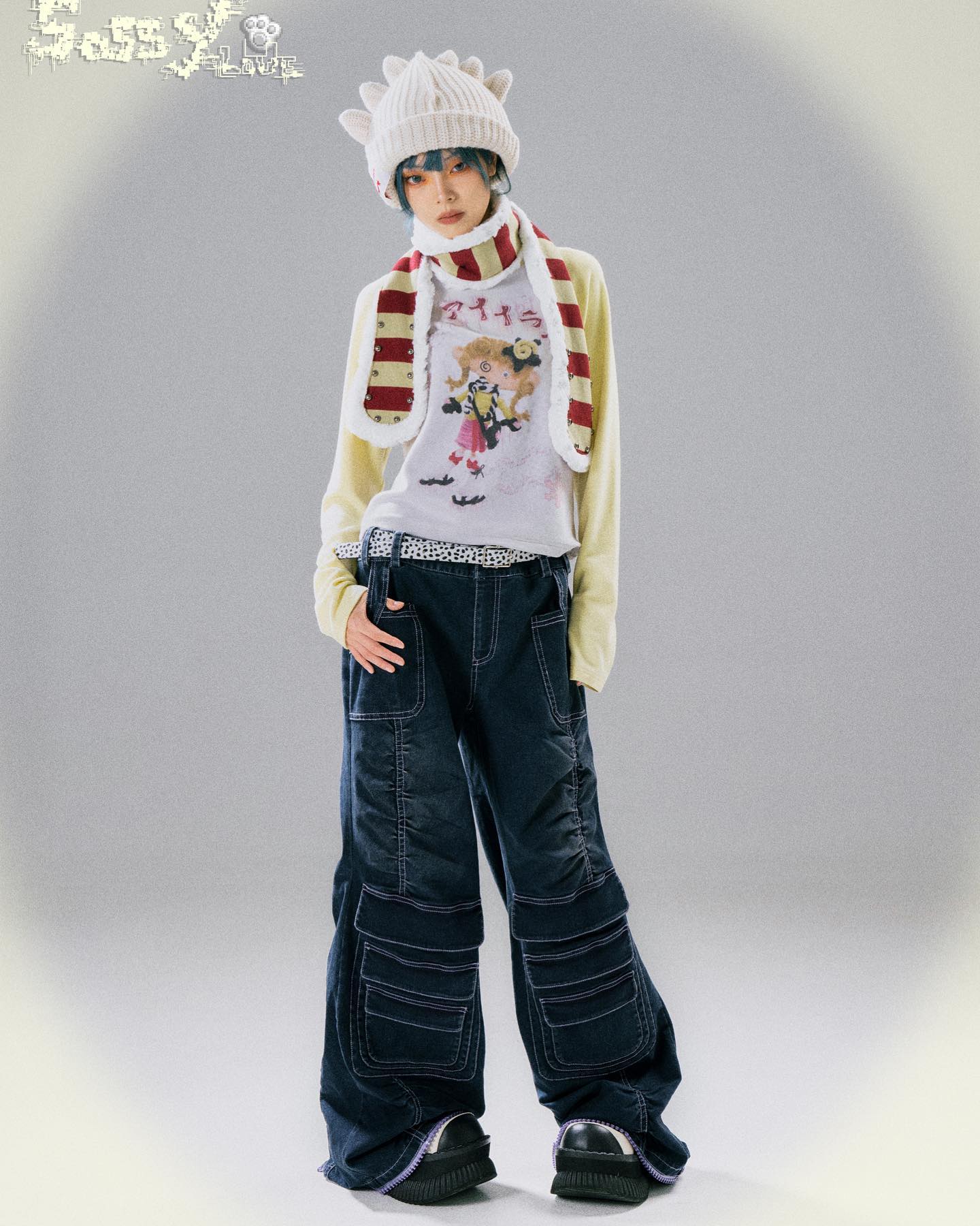 a full shot of a model wearing a complete outfit. the model is wearing a white knit beanie with spikes on top, a creamcolored longsleeved shirt with a graphic tee underneath featuring an anime girl, a red and cream striped scarf, darkwash wideleg cargo pants with multiple pockets and stitching details and platform shoes. the backdrop is simple, providing focus on the model and outfit. the overall style is a mix of streetwear, gothic lolita and harajuku fashion.
