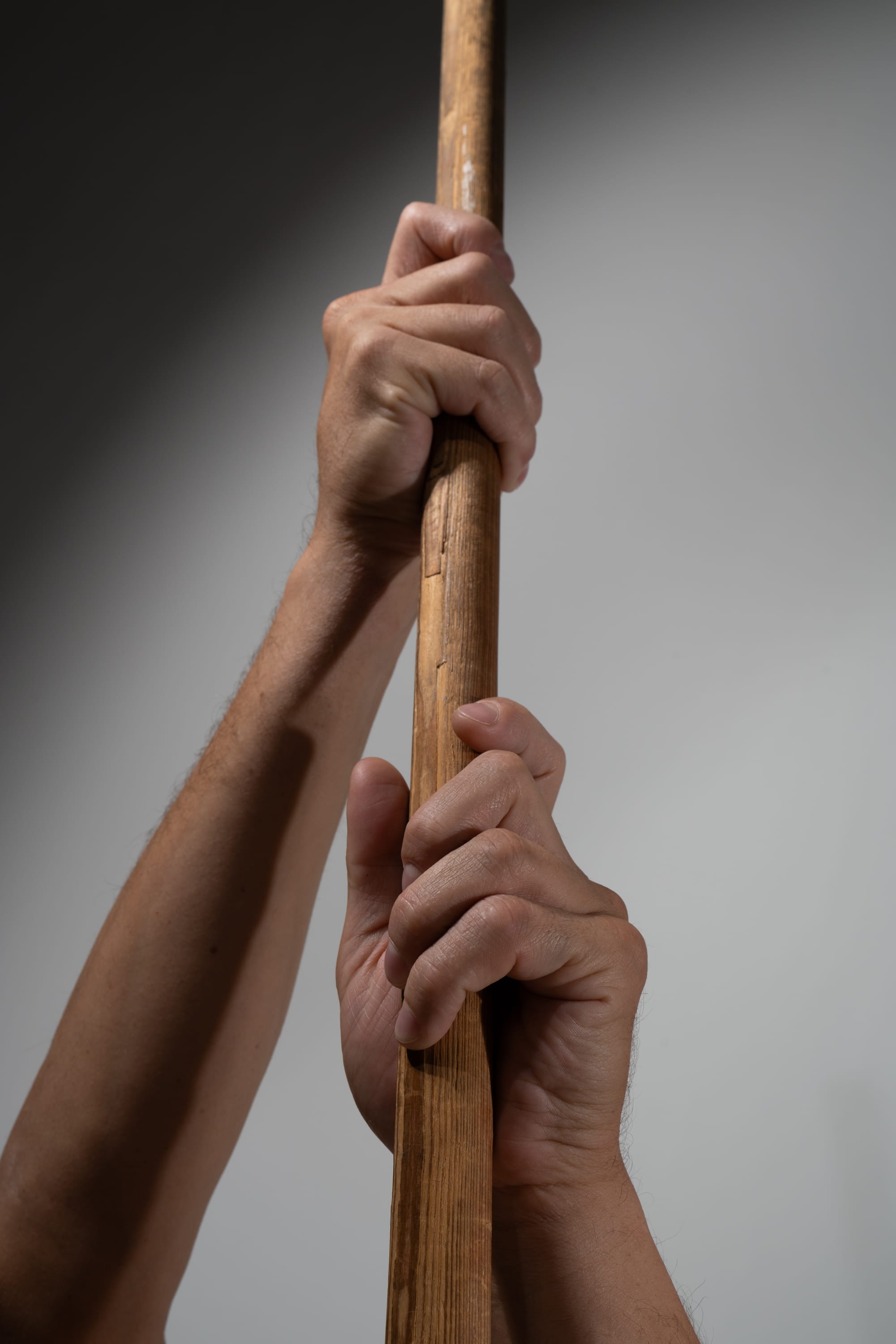 the image shows a pair of hands tightly gripping a roughhewn wooden pole. the pole is positioned vertically, and the hands are holding it firmly as if in the midst of a strenuous activity.  the lighting is dramatic, casting shadows that emphasize the tension and effort in the hands.
