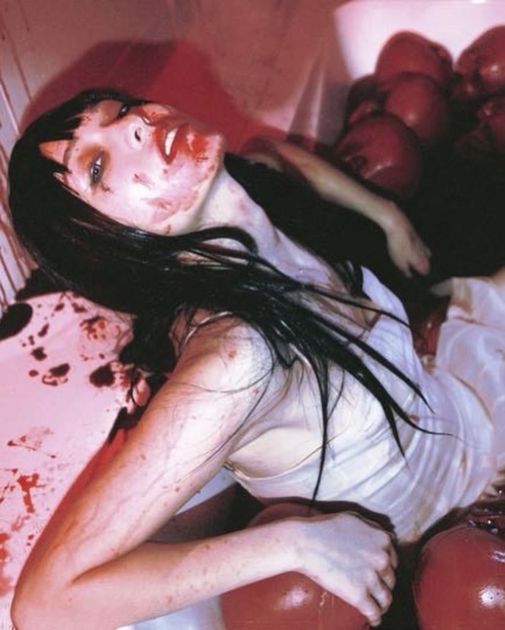 a woman is in a bathtub filled with red balloons and what appears to be blood. she is covered in blood, especially around her face, and also has bruises on her arm. the scene is disturbing and unsettling.