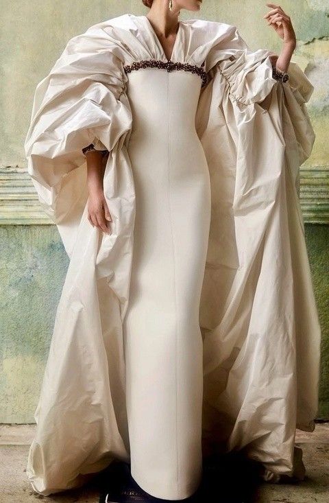 a woman is wearing a long, creamcolored gown with voluminous, dramatic sleeves. the dress has a simple, fitted silhouette, while the sleeves are oversized and gathered. there is embellishment detailing around the neckline of the dress. the overall look is elegant and fashionable.