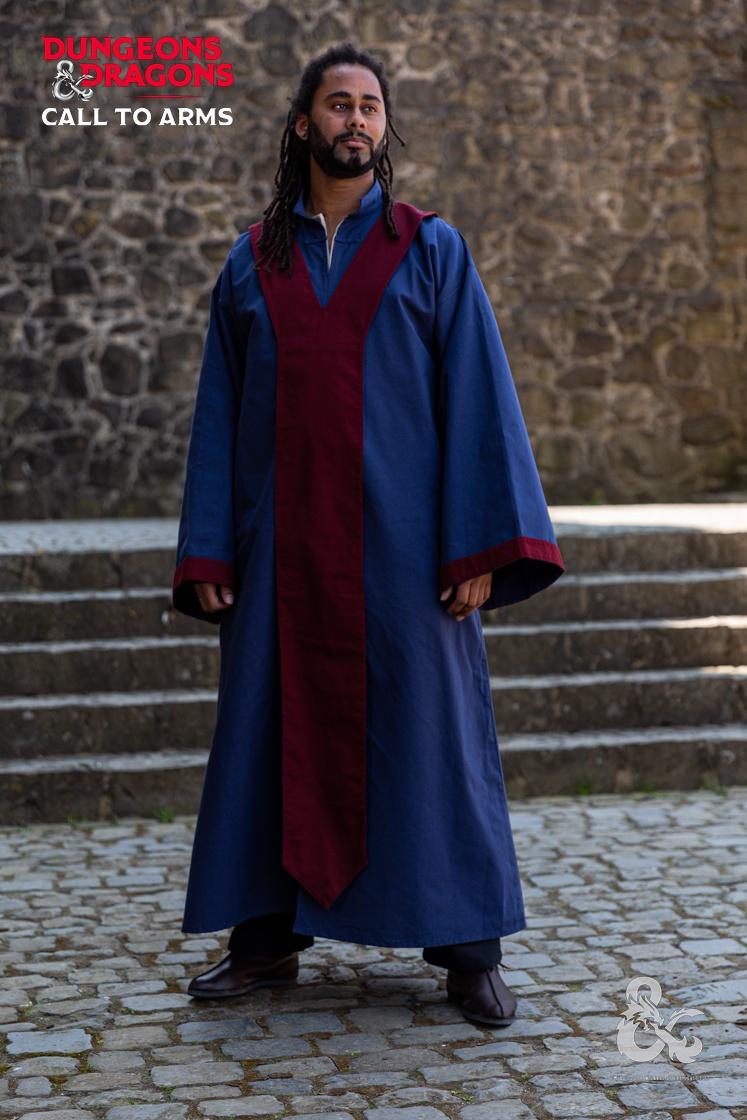 a man with dreadlocks and a beard stands outdoors, wearing a long blue robe with a maroon vertical stripe down the front. he is standing on a cobblestone surface, with stone steps and a stone wall in the background. the dungeons & dragons "call to arms" logo is in the upper left corner and the lower right corner.