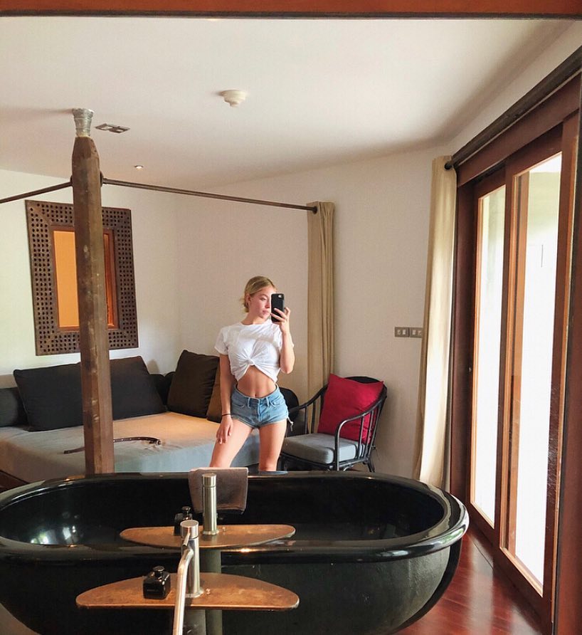 a blonde woman takes a selfie in a luxurious hotel bathroom.  she's wearing a white knotted shirt and denim shorts and is standing in front of a large black freestanding bathtub. the bathroom is stylishly decorated with a modern minimalist feel. the room features a fourposter bed visible in the background, wooden floors, and large windows that let in natural light. the overall ambiance is one of relaxation and highend travel.