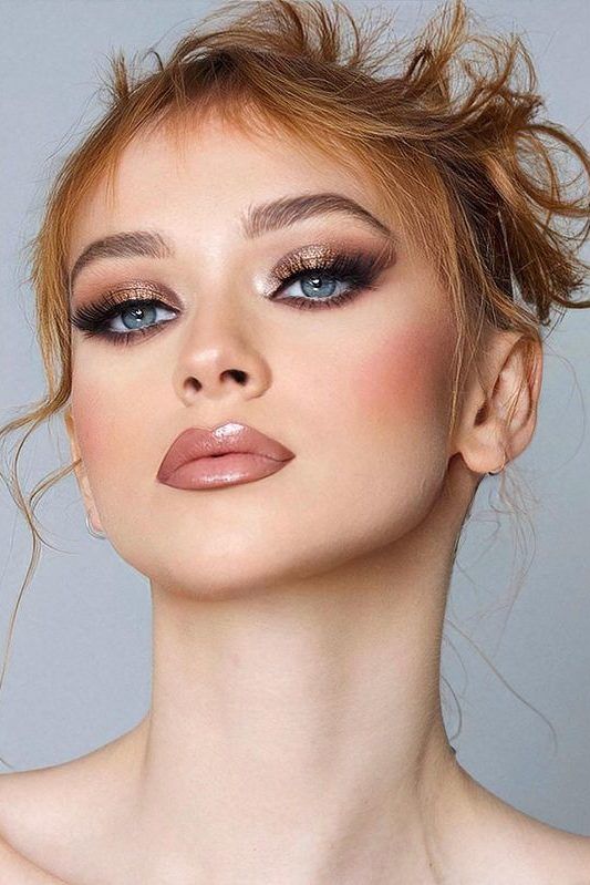 a closeup portrait of a woman with a beautiful makeup look. she has light skin, rosy cheeks, and blue eyes. her lips are covered in nude lipstick with a glossy finish. she is wearing a gold and black eyeshadow with dramatic false eyelashes. her eyebrows are welldefined. her orangeblonde hair is styled in a messy updo. she is facing the camera. the image is a studio shot with soft lighting. the image has a glamorous and elegant feel.
