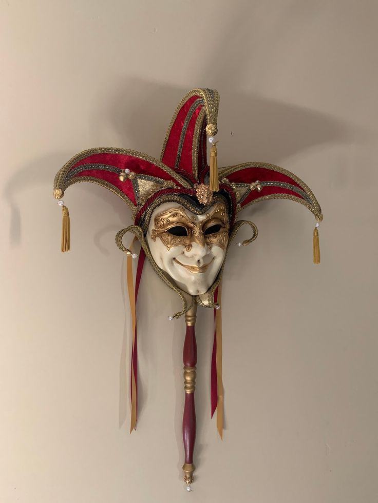 a decorative venetian jester mask hangs on a wall. the mask is mounted on a handle, and features a red and gold jester hat with tassels and beads. the mask itself is white with gold detailing around the eyes and nose. ribbons hang from the base of the mask. the handle is red with gold accents.
