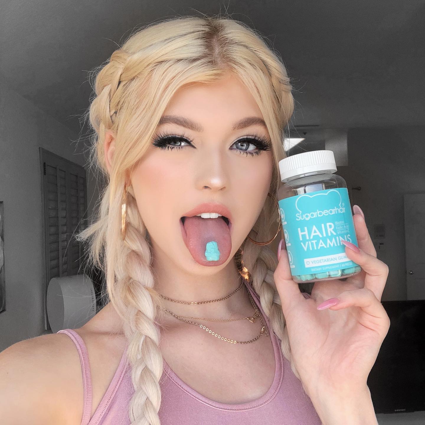a blonde woman, likely loren gray, is posing with a bottle of sugarbearhair hair vitamins and a blue gummy bear on her tongue. she has braided hair, wears gold hoop earrings and layered necklaces, and is dressed in a pink tank top. the image appears to be a product promotion or endorsement.