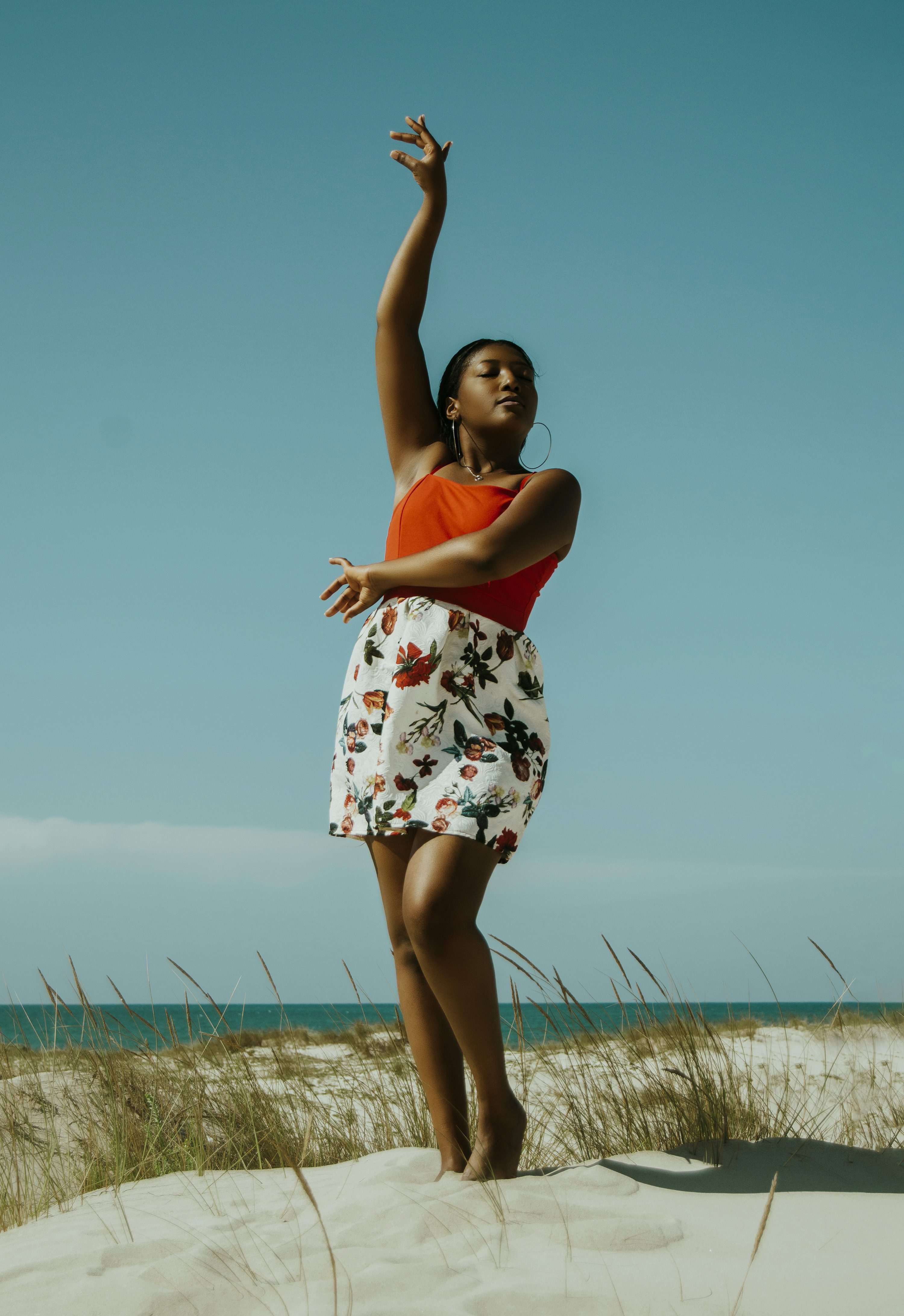 the image shows a black woman standing gracefully on a sandy beach against a vibrant blue sky. she is wearing a short, floral print skirt and a bright orange top.  one arm is raised elegantly, suggesting a dancelike pose. the overall mood is one of freedom, joy, and expressive movement, enhanced by the bright sunlight and natural setting.  the woman's posture and expression convey a sense of confidence and poise.