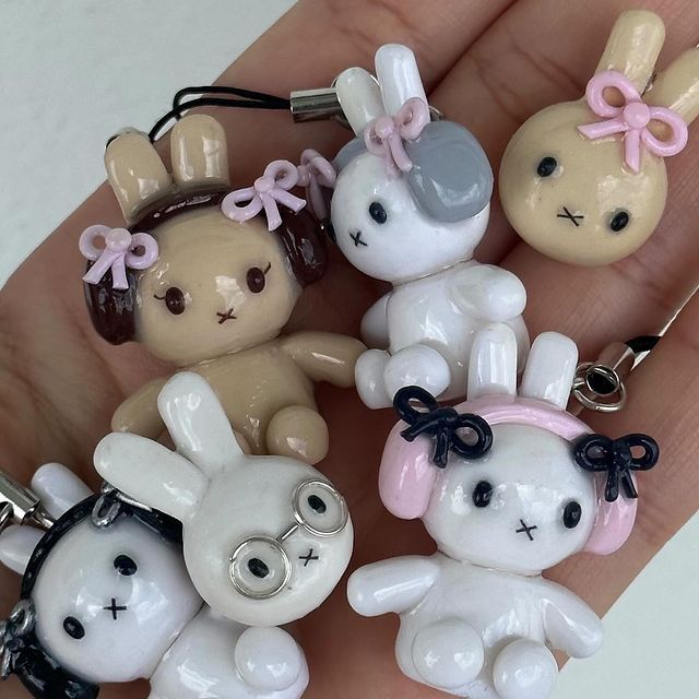 the image shows a hand holding several small, handmade bunny figurines. the figurines are made of clay or a similar material and are decorated with bows, glasses, and other accessories. the figurines are various colors, including pink, white, gray, brown, and black. they are kawaiistyle, meaning they have a cute and cartoonish appearance. they appear to be attached to phone charms.