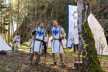 picture of two knights in medieval armor standing in a forest setting near tents. they are wearing white and blue armor with an eagle emblem. they each carry a type of pole weapon.  other figures are visible in the background, seemingly part of the same group. the setting suggests a historical reenactment or a depiction of a medieval camp.  a banner with a blue and white design stands behind the central figures.