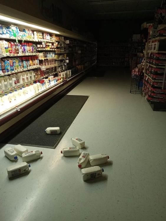 the image shows the dairy aisle of a grocery store with multiple gallons of milk spilled on the floor. the aisle is dimly lit and the shelves are stocked with various dairy products. there are no people in the image.