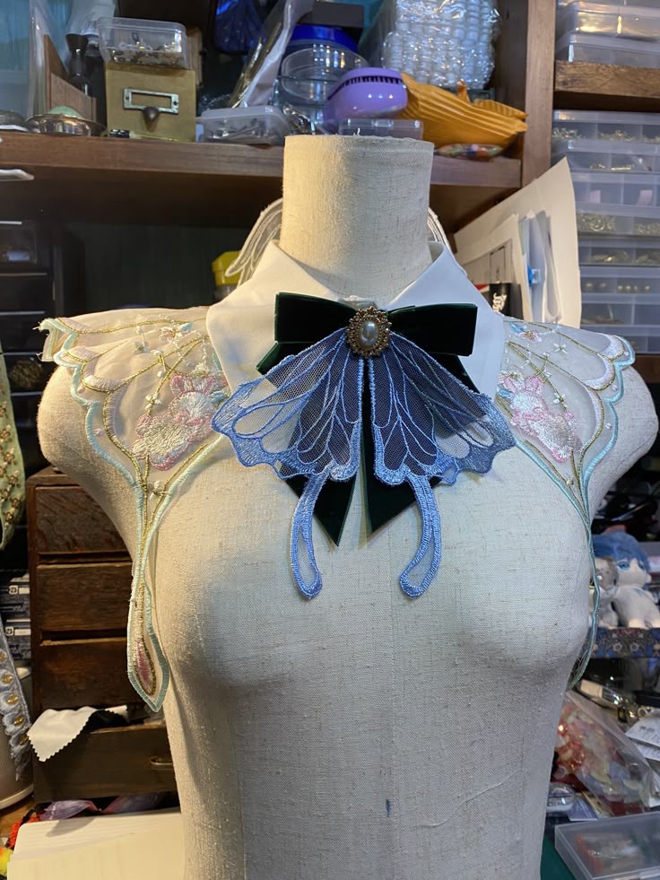 a mannequin is adorned with a white collar, a green velvet bow with a decorative brooch, and intricate embroidered lace shoulder pieces with floral and butterfly designs. the scene suggests a crafting or sewing setting.