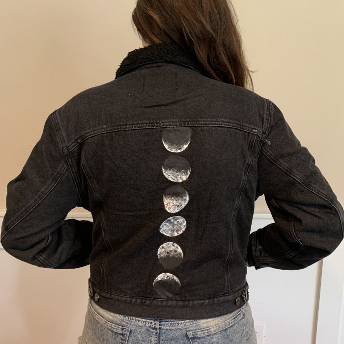 a woman is modeling a black denim jacket with a design of moon phases painted on the back. the photo is taken from behind, showcasing the design.