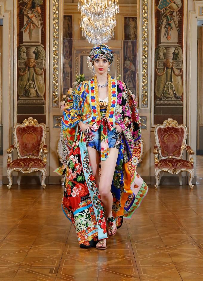 a female fashion model walks the runway wearing a colorful patterned  dress with a  gold chain necklace and sandals on her feet. she is wearing a headscarf and has  gold jewelry on her fingers. the background features two gold and red antique chairs and a wooden parquet floor. the walls are adorned with elaborate ornate paintings. the image is likely from a highfashion runway show.