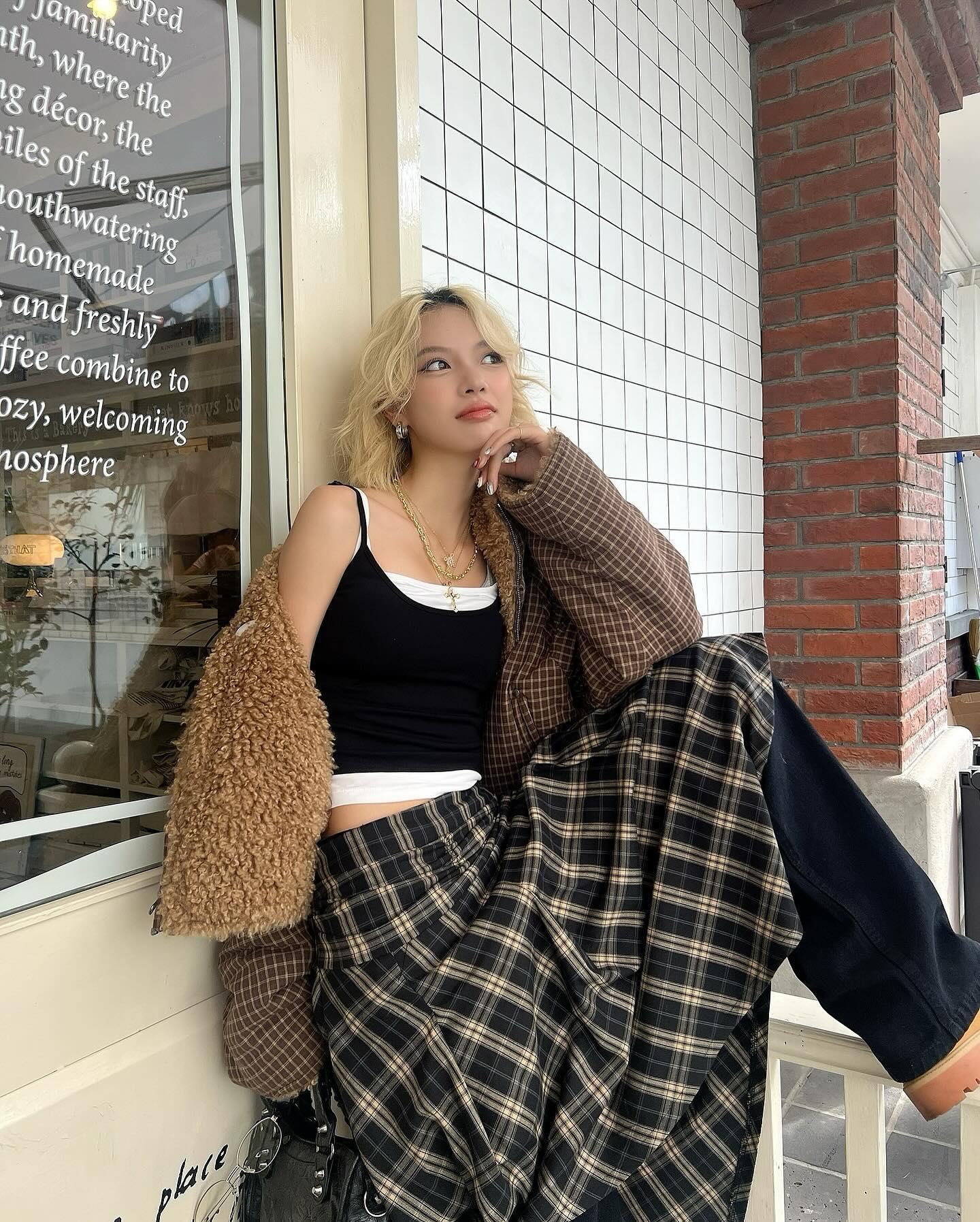 a young blonde woman is standing outside a cafe.  she's wearing a black tank top, a brown plaid maxi skirt, a light brown teddystyle jacket, and black pants underneath the skirt.  the cafe's window displays text about its cozy atmosphere and features. she's posing casually, looking slightly away from the camera. the overall style is stylish and leans towards retro or vintage aesthetics.