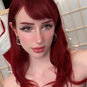 closeup portrait of a young, attractive woman with long, wavy, bright red hair. she's wearing makeup that includes winged eyeliner and glossy pink lipstick.  she has on clear hoop earrings.  her shoulders are visible, and part of a christmas stocking is seen in the background. the background is softly lit and features a lightcolored screen or room divider. the overall impression is a stylish, selftaken portrait with a slightly festive feel.