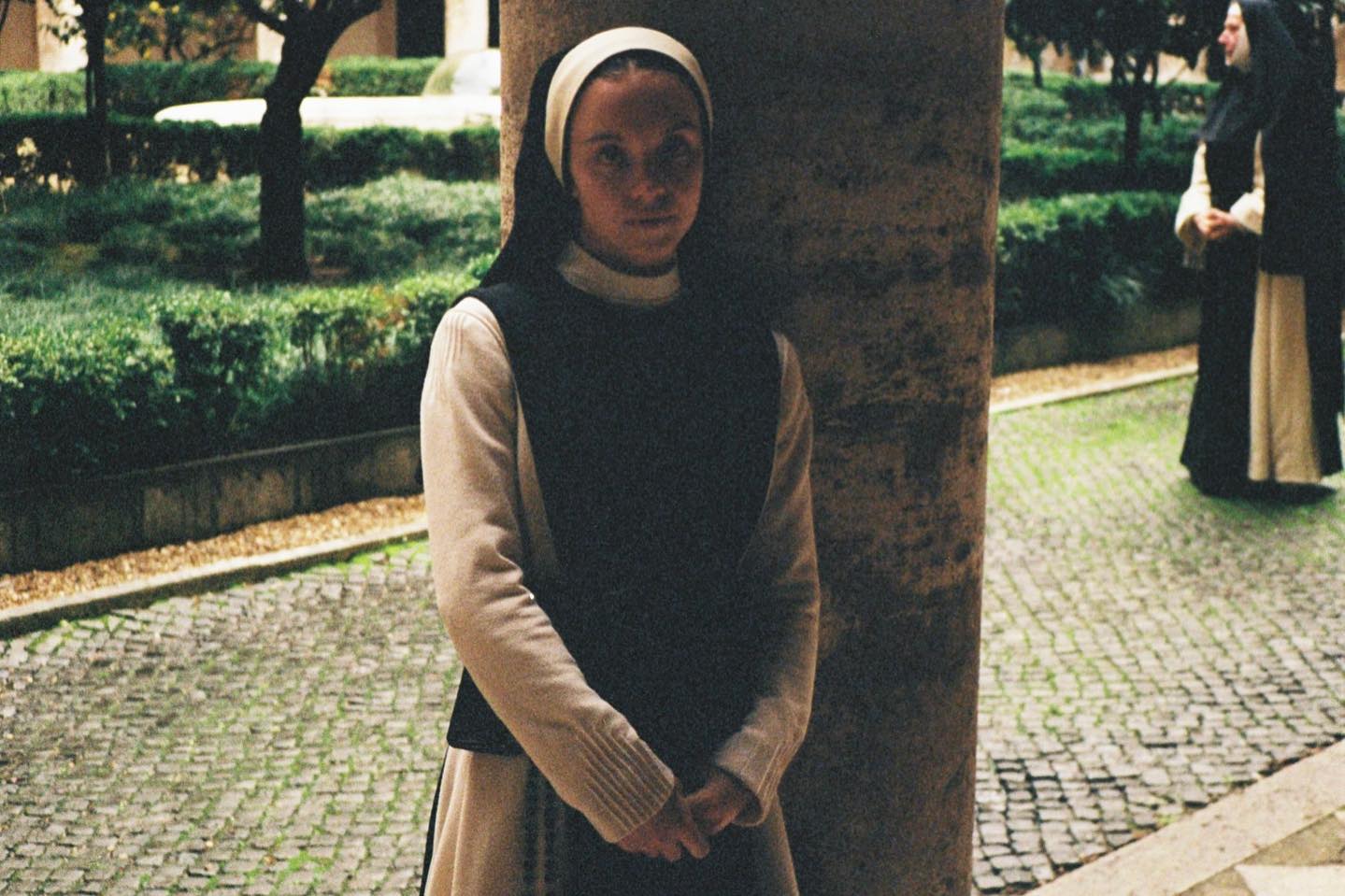 the image shows a young nun in a beige and dark habit standing close to a stone pillar in a monastery courtyard.  the courtyard features a neatly manicured garden with hedges and stone pathways. another, older nun in a darker habit is visible in the background, further away. the overall mood is serene and contemplative. the lighting is somewhat muted, consistent with an overcast day or indoor lighting. the focus is primarily on the young nun in the foreground.