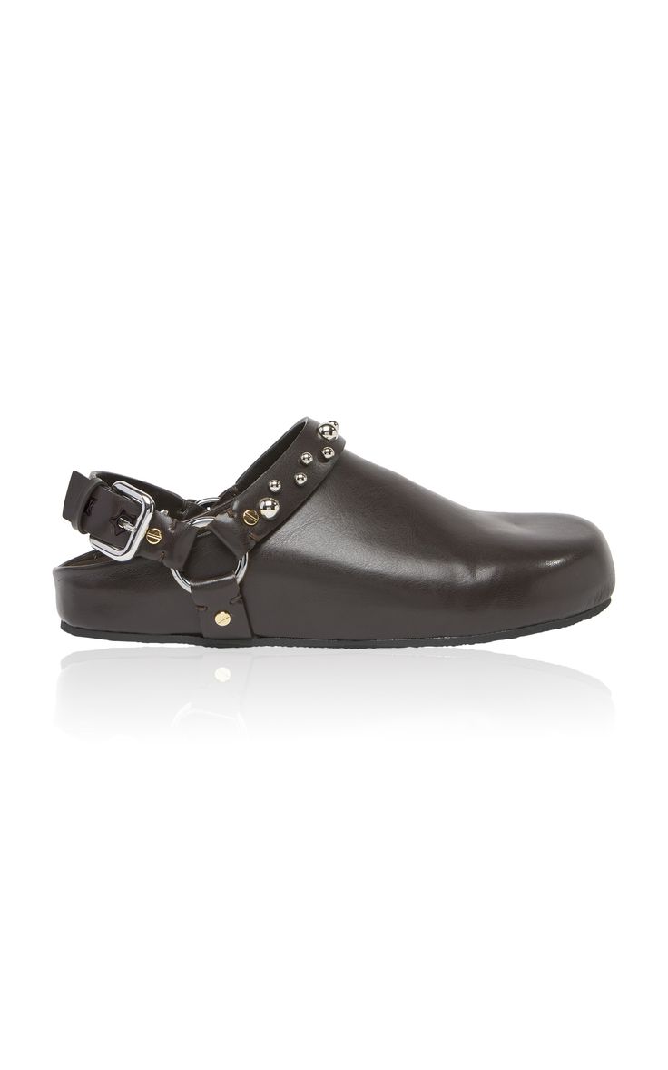 pair of dark brown leather clogs with silver studs and buckles are presented against a white background. the clogs have a slightly raised platform sole and a simple, elegant design. the image is welllit and shows the shoes in detail, highlighting their texture and features.
