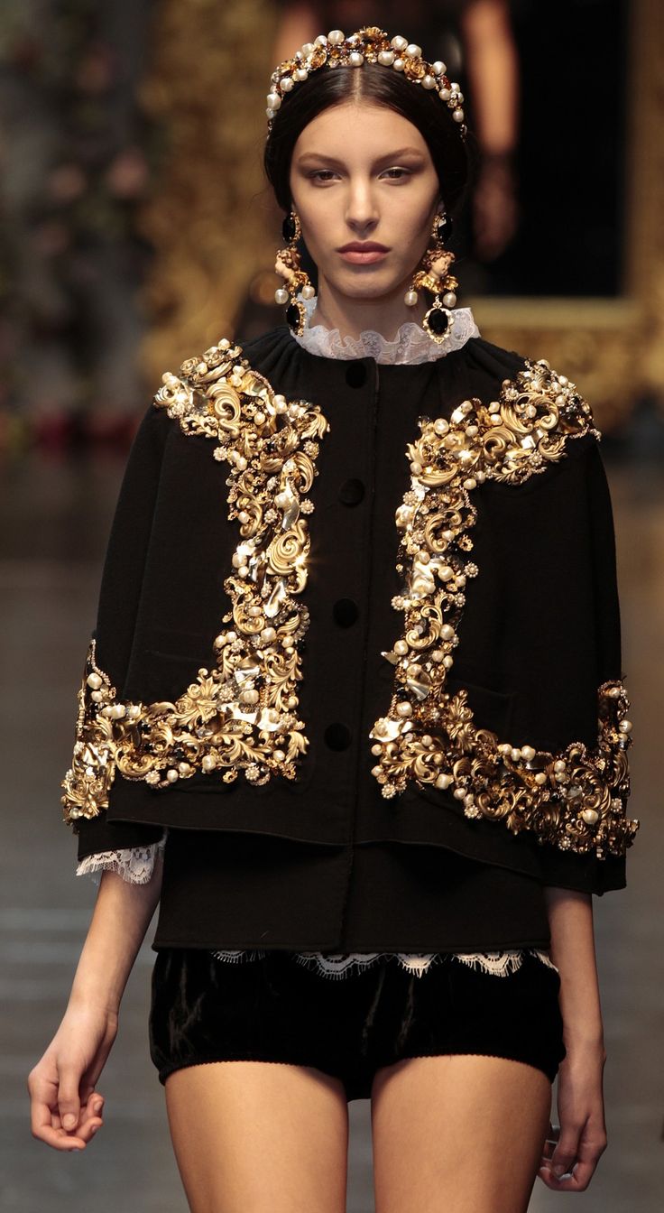 a fashion model walks the runway wearing a stunning dolce & gabbana outfit.  the outfit consists of a black bolero jacket heavily embellished with gold and pearl detailing, a black velvet miniskirt with a lace trim, and intricate gold and pearl earrings and a matching headband. the model's makeup is subtle and elegant, highlighting her natural features. the background is blurred, focusing attention on the model and her outfit. the overall image projects luxury, elegance, and high fashion.