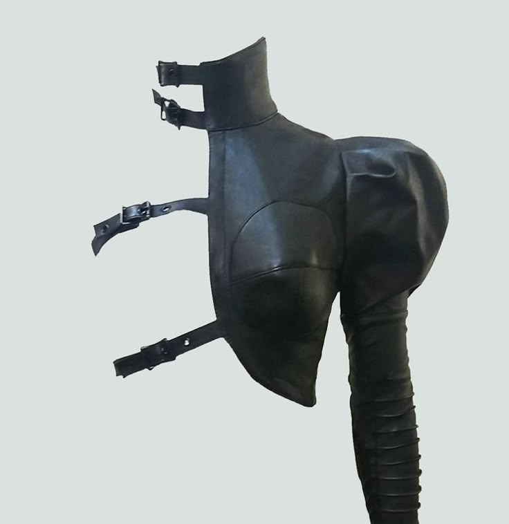 a black leather garment with a high collar and multiple straps with buckles is displayed against a light gray background. the garment features a fitted upper section with a padded or bulbous detail at the shoulder, leading into a long, fitted sleeve with horizontal textured lines.
