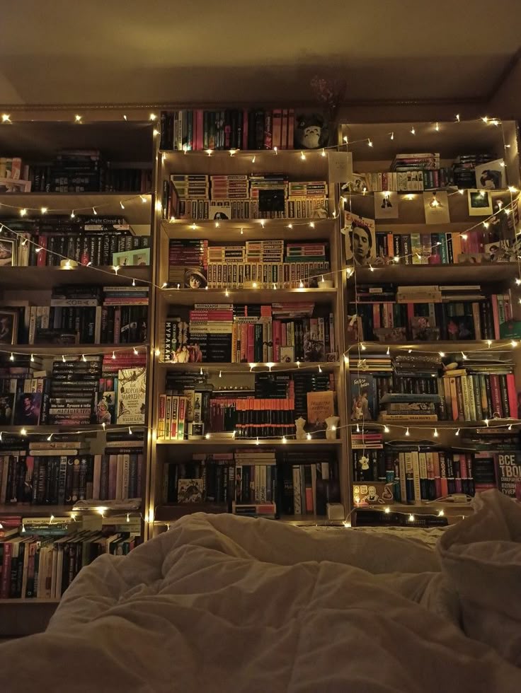this image shows a cozy bedroom interior featuring three bookshelves filled with books and decorated with fairy lights. in the foreground, there is a bed with white bedding. the warm light from the fairy lights adds to the cozy atmosphere of the room.