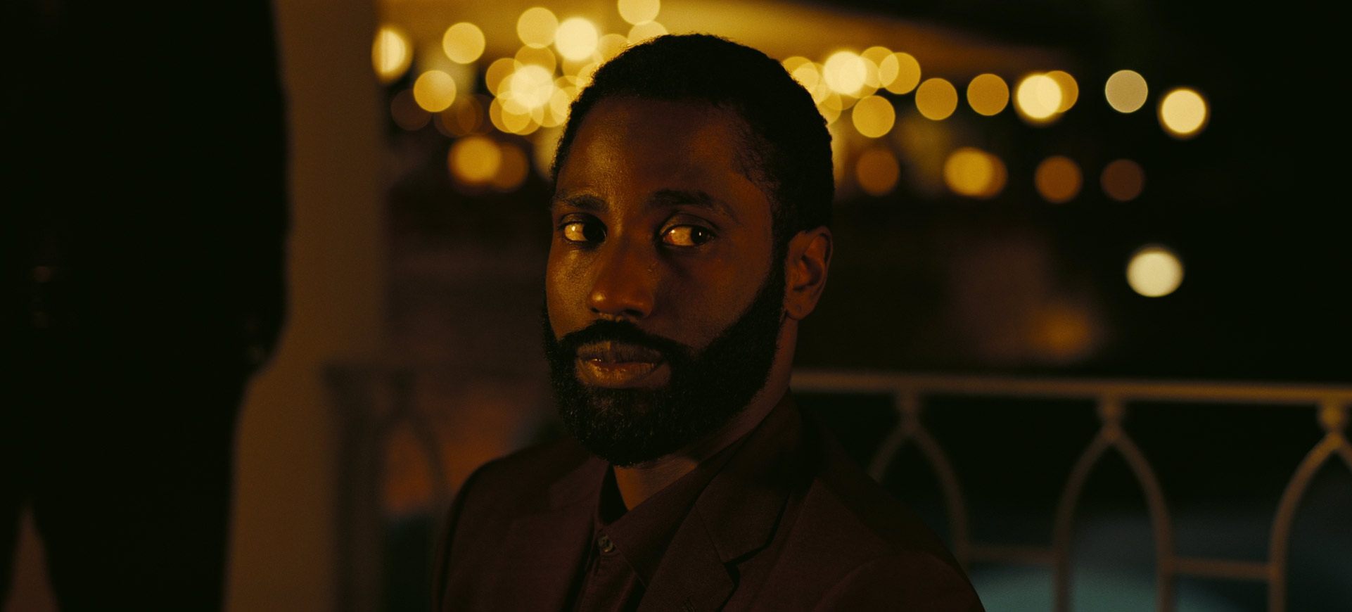 a dark, moody image of a black man looking directly at the camera.  he has a serious expression on his face and his dark eyes are piercing. he is wearing a suit jacket and his hair is closely cropped. the background is blurred with bokeh lights in the distance.  the image is lit by warm, yellow light and has a film noir vibe.