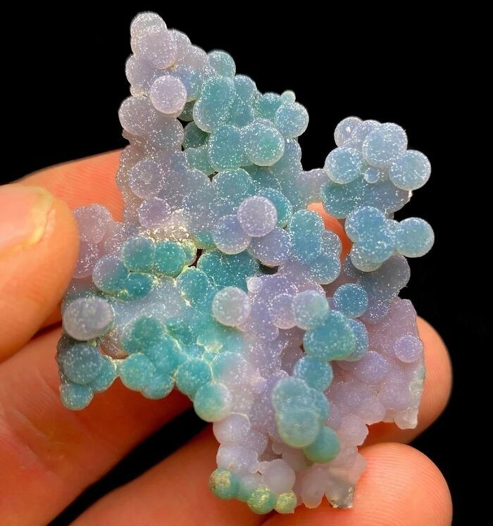a closeup shot of a beautiful, polished agate specimen. the agate is composed of many small, round, iridescent nodules that have a range of colors, including purple, green, and blue. the agate is held in a person's hand, showcasing its size and intricate detail. the background is black, providing good contrast for the vibrant agate. the overall image is a captivating display of nature's artistry and the beauty of gemstones.