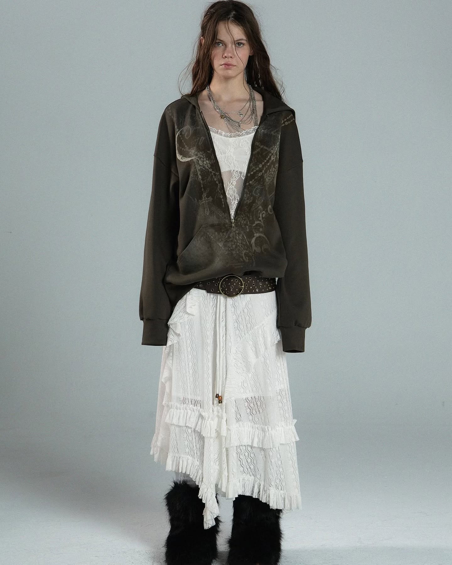 a fulllength shot of a female model showcasing a stylish outfit. she's wearing a dark olive green oversized hoodie with a unique, possibly tiedye or distressed, pattern. underneath, she has on a white lacy, tiered midi skirt with a ruffled asymmetrical hemline.  the model also sports a delicate layered necklace, a brown belt, and black furry boots. her pose is straightforward, and the background is a plain light grey, keeping the focus on the clothing. the overall aesthetic is a blend of bohemian, grunge, and vintage elements.