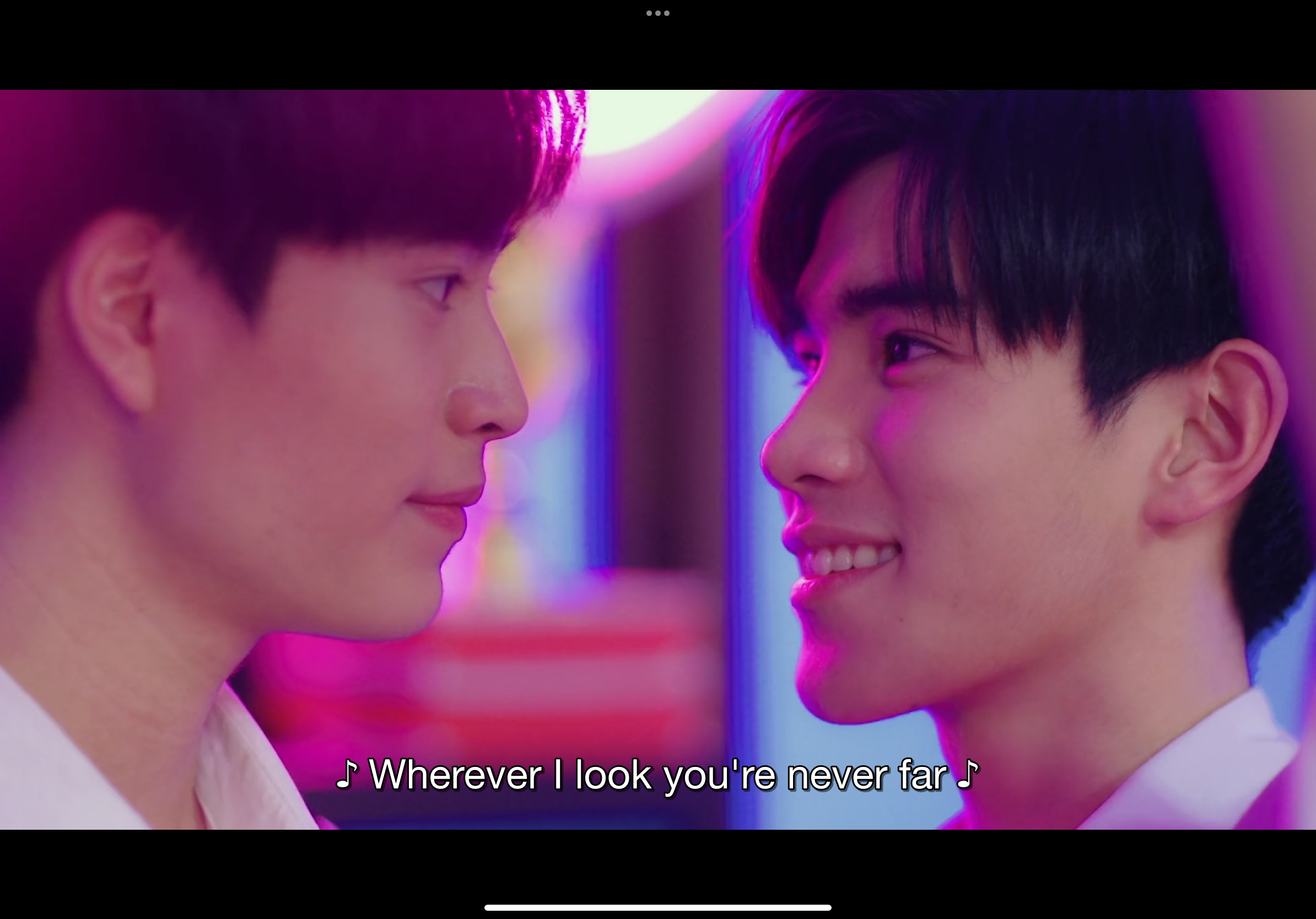 the image shows two young men gazing at each other in a dimly lit space illuminated by neon lights. they are positioned very close to one another and both have soft smiles on their faces, suggesting an intimate and affectionate relationship. the man on the left wears a white shirt while the man on the right wears a black shirt.  the text overlay reads "wherever i look you're never far" implying a romantic context.