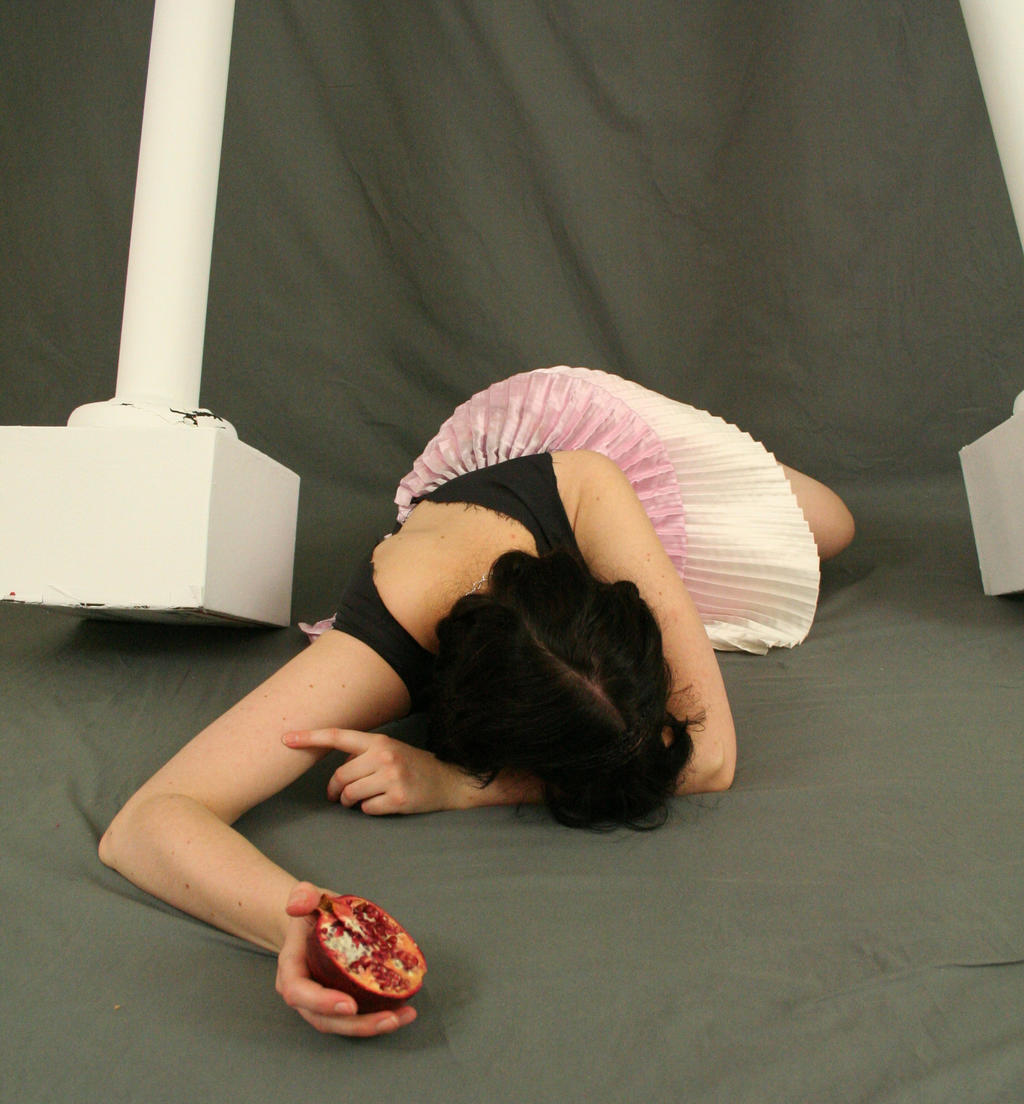 the image shows a young woman lying on a gray floor, her body angled slightly away from the viewer. she's wearing a black top and a pink and white pleated skirt, which resembles a tutu.  her dark hair is spread out around her head. one arm is outstretched towards a half of a pomegranate that rests near her hand. the background features two partially visible white columns. the overall mood is artistic and somewhat dramatic. the lighting and composition suggest a posed studio photograph.