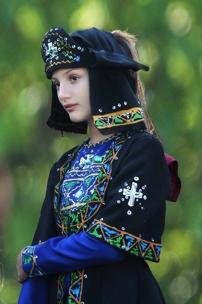 a young girl in traditional clothing, including a black headdress with embroidery and a cross symbol, a blue and green top with similar embroidery, and a black jacket with matching details. she stands against a blurry green background.