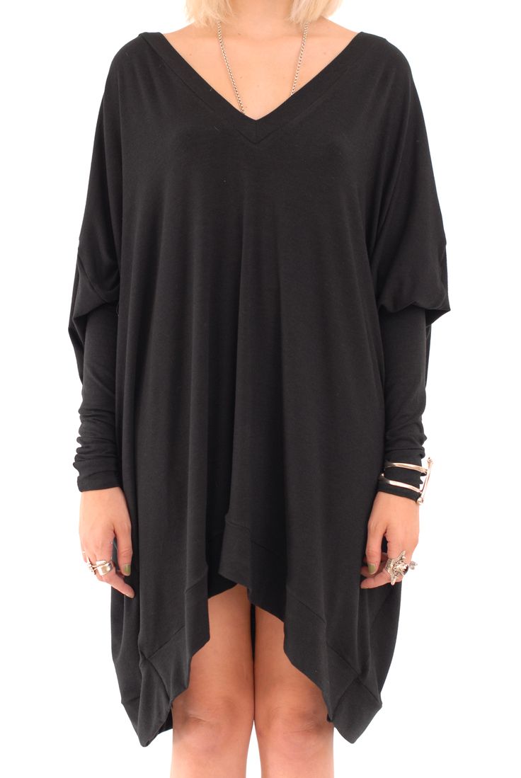 a person is wearing a loosefitting, longsleeved black tunic dress with a vneck. the dress is an oversized style and features an asymmetrical hemline. the person is wearing jewelry, including rings and a bracelet. a chain necklace is also visible. the image is against a plain white background.