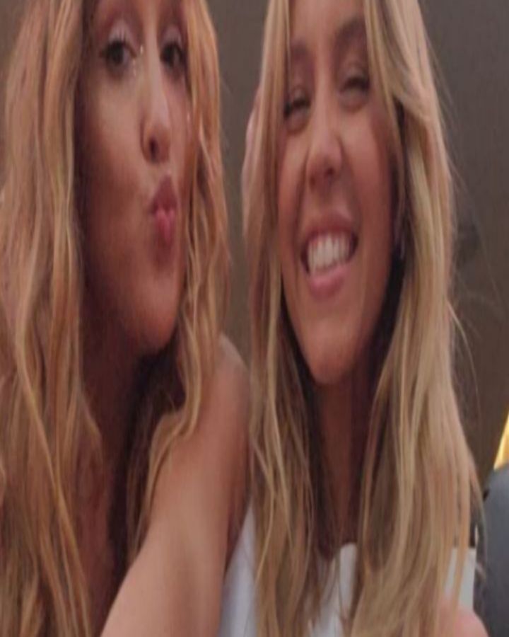 a closeup shot of two young women with long, wavy blonde hair.  they are close together, one woman has her arm around the other, and they appear to be taking a selfie. both are smiling; one is making a kissy face, the other has a wide, happy smile.  they look like friends or sisters. the setting appears to be indoors, and the lighting is soft. the overall mood is warm, friendly, and intimate.