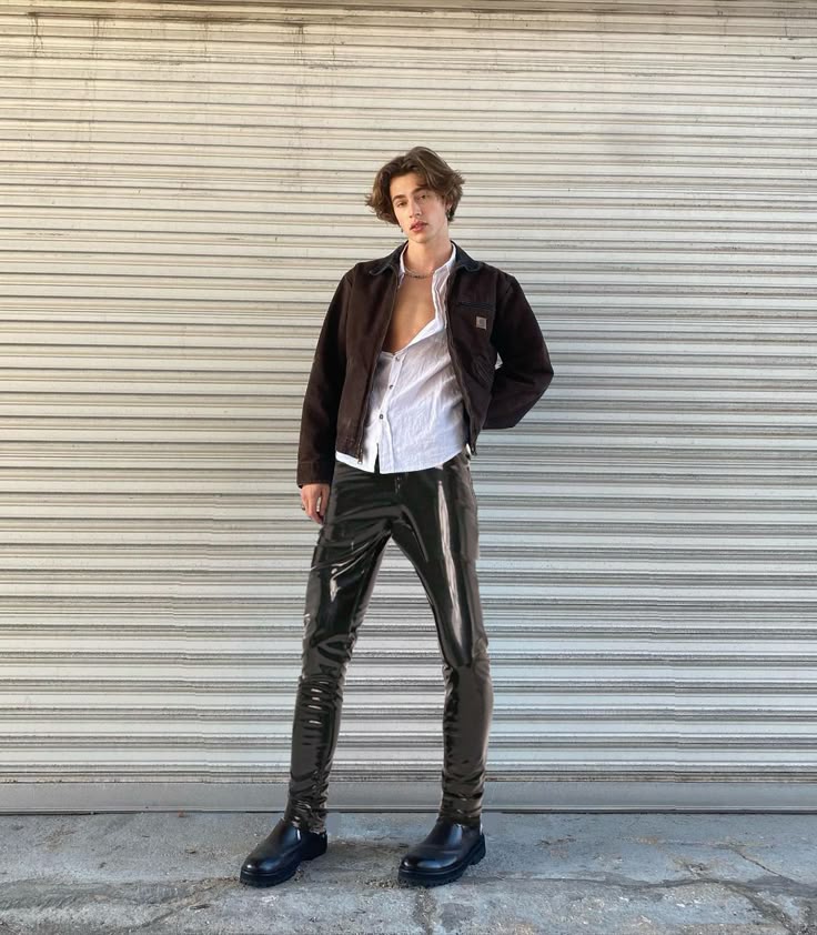 a young man stands confidently in front of a corrugated metal wall. he is wearing a brown jacket over an unbuttoned white shirt, paired with shiny black pants and black shoes. his casual yet stylish outfit and pose suggest a fashion or urban setting.