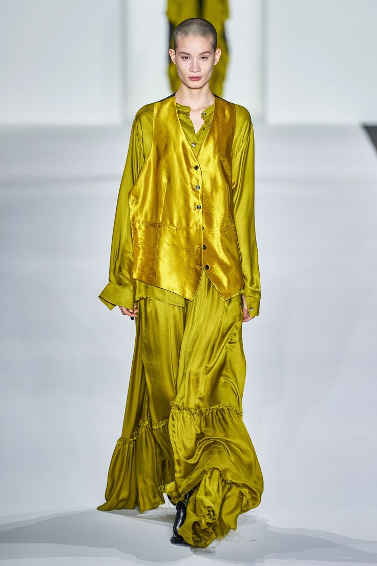 a fashion model walks down the runway wearing a long yellow silk dress with a satin vest and black shoes. she has a bald head and is looking straight ahead. the model is posing for the camera, and the dress is flowing beautifully as she walks. the runway is white and the background is blurred. the model is wearing stylish, elegant clothing. the dress is very chic and formal. this is a high fashion runway show. the dress is contemporary and very stylish.