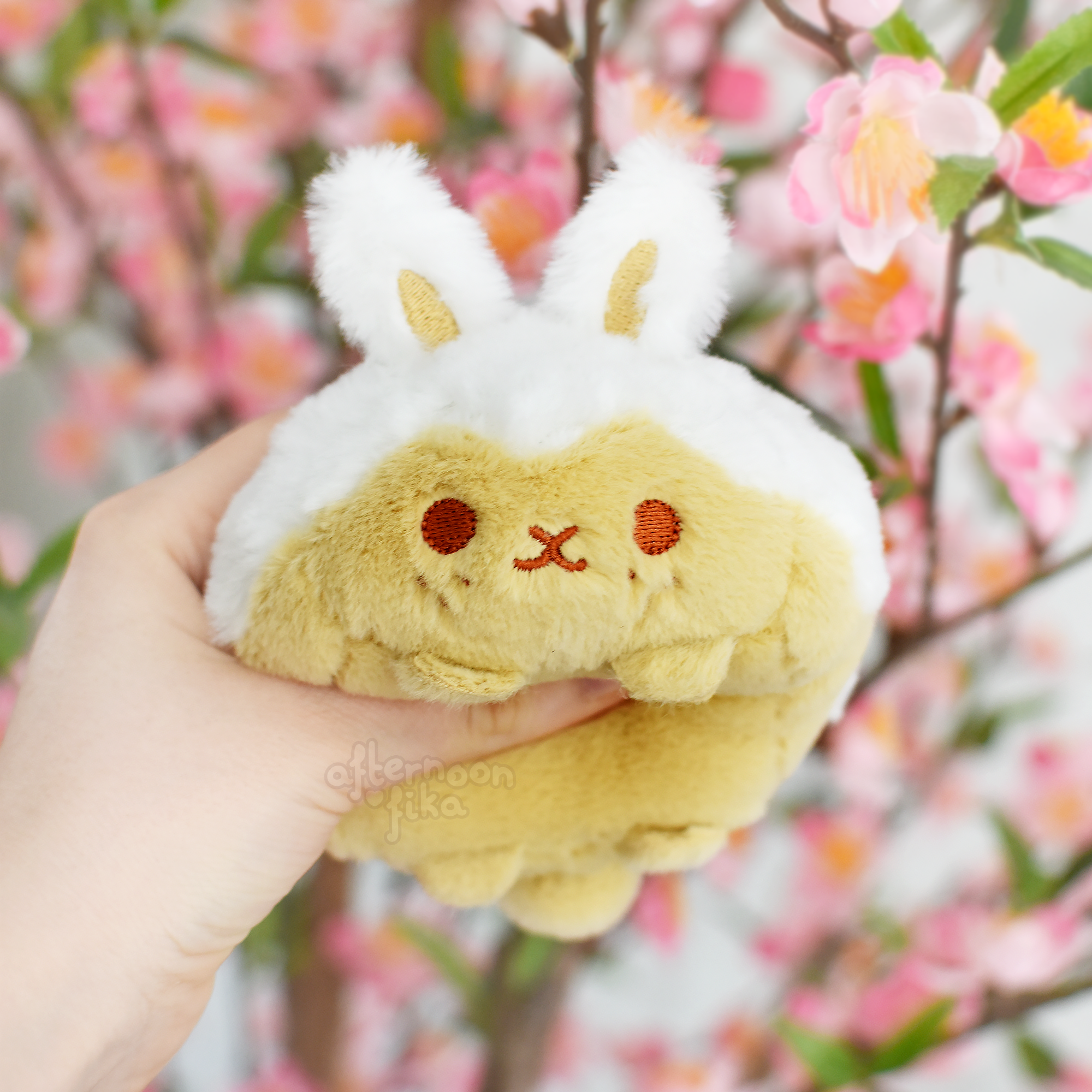 a closeup shot of a hand holding a cute plush toy bunny. the bunny is primarily light yellow/tan with white ears and features a sweet, simple facial expression. the background is blurred but showcases a blossoming cherry tree with pink flowers, creating a soft and charming atmosphere. the plush toy appears soft and huggable, and the overall image evokes feelings of warmth and cuteness.