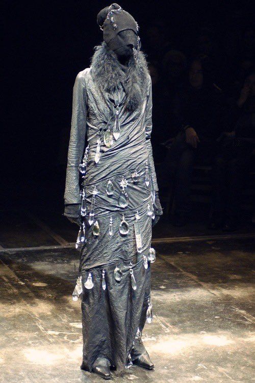 a fulllength shot of a model on a runway, wearing a darkcolored, possibly black or deep gray, dress and headwear that obscures their face. the dress appears to be of an avantgarde or deconstructed design, featuring intricate draping and a textured fabric. the model is also wearing a fur collar. crystallike accessories or jewelry are attached to the dress. the lighting is dim and the background is dark, focusing attention on the model and the unique fashion ensemble. the setting is a typical fashion show, likely on a catwalk with an audience in the background, though the background is mostly obscured by shadow.