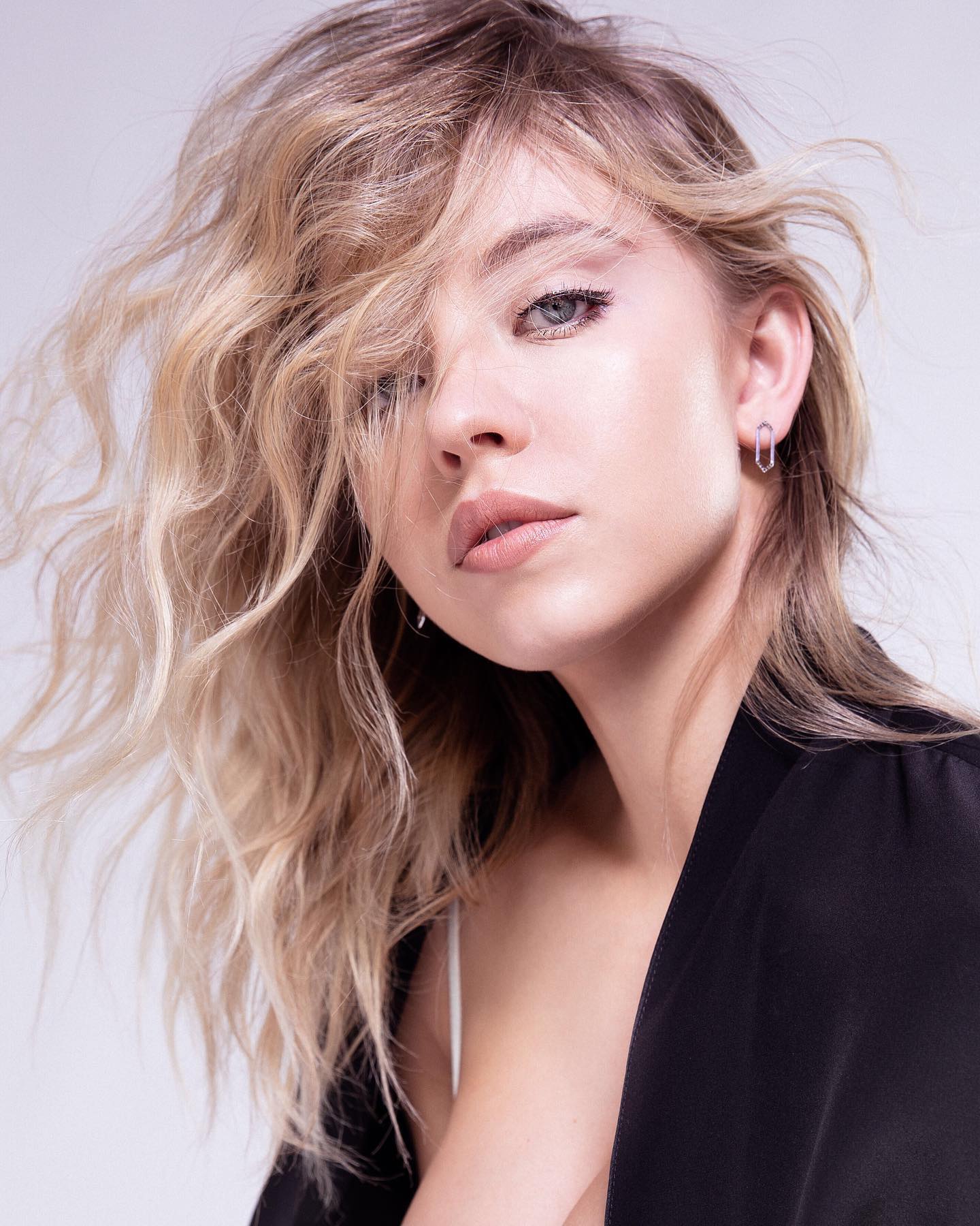 a closeup portrait of a young, attractive blonde woman with voluminous, wavy hair.  her hair partially obscures her face, adding a touch of mystery. she's wearing a black blazer, partially open, revealing a glimpse of a white undergarment. her makeup is subtly done, emphasizing her eyes and lips. the background is plain and light, keeping the focus on the model. the overall impression is one of casual elegance and beauty.
