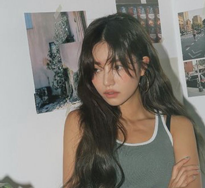 a beautiful asian woman with long black hair, wearing a gray tank top is looking at the camera with a light smile. the background has some pictures behind her.