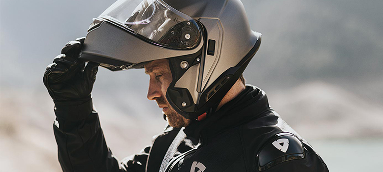 a motorcyclist wearing a black leather jacket, a gray helmet, and gloves. he is looking down with his visor open. the background is blurry, but it looks like a mountain range or a rocky area.  the motorcyclist is wearing protective gear and is ready for a ride.