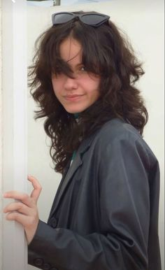 a young woman with long, curly brown hair is wearing black sunglasses and a black leather jacket. she is smiling and looking at the camera. the photo is a closeup and is taken from the front.