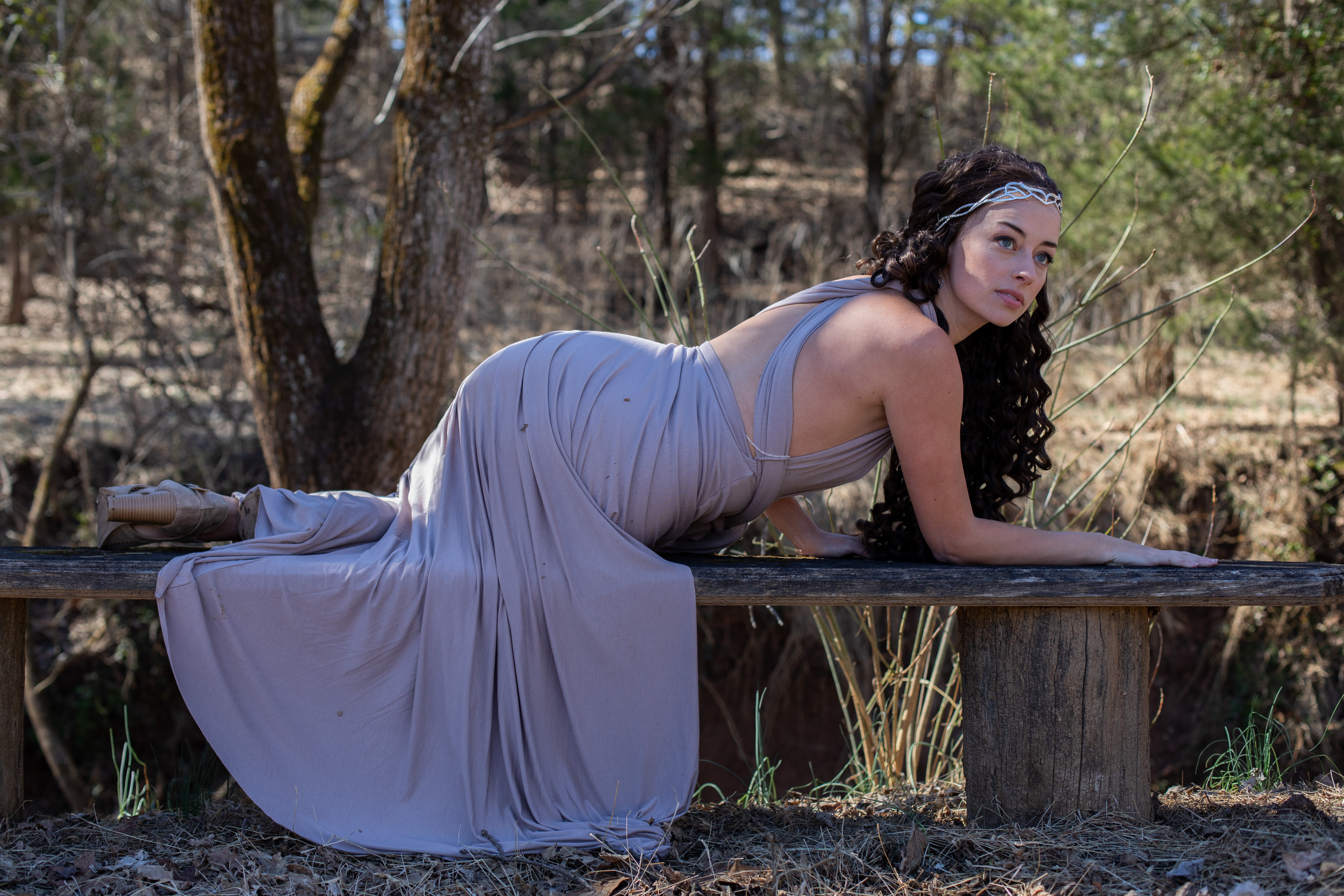 a woman in a long flowing lavender dress poses on a wooden bench in the forest. she has long dark curly hair and is wearing a silver headband. the photo is taken from eye level with natural lighting. the environment is outdoors and the perspective is in focus.