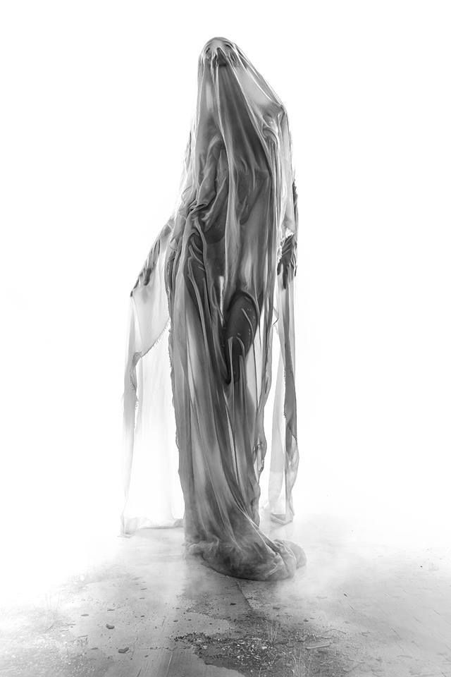 the image is a black and white, fullbody shot of a woman seemingly enveloped in a large, sheer, translucent fabric. the fabric drapes around her body in a way that obscures her features and form, giving a ghostly or ethereal appearance.  the fabric's texture is visible, showing folds and creases. the woman appears to be standing on a rough, textured floor. the overall feel is mysterious and artistic, possibly conceptual in nature, with high contrast between the light and dark areas of the image. the background is plain white, emphasizing the subject.