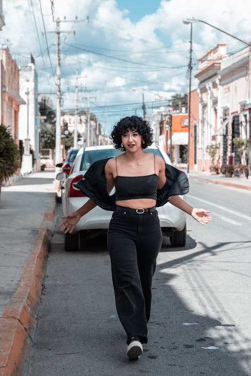 a young latina woman with short curly hair is walking down a city street. she is wearing a black crop top, wideleg black pants, and black sneakers. she is carrying a black jacket over her shoulder and has a happy and confident expression on her face. the street is lined with colorful buildings and parked cars. the sun is shining brightly.