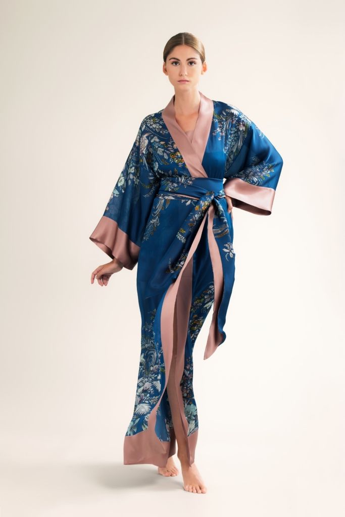 a fulllength shot of a woman model showcasing a luxurious blue and pink floral silk kimono robe. the robe is long, with wide sleeves and a wrap closure, tied at the waist with a belt.  the model is standing with a confident pose against a plain light beige background.  the kimono has a teal blue base with a pink underlining and detailed floral pattern throughout. the overall impression is one of elegance and sophistication.