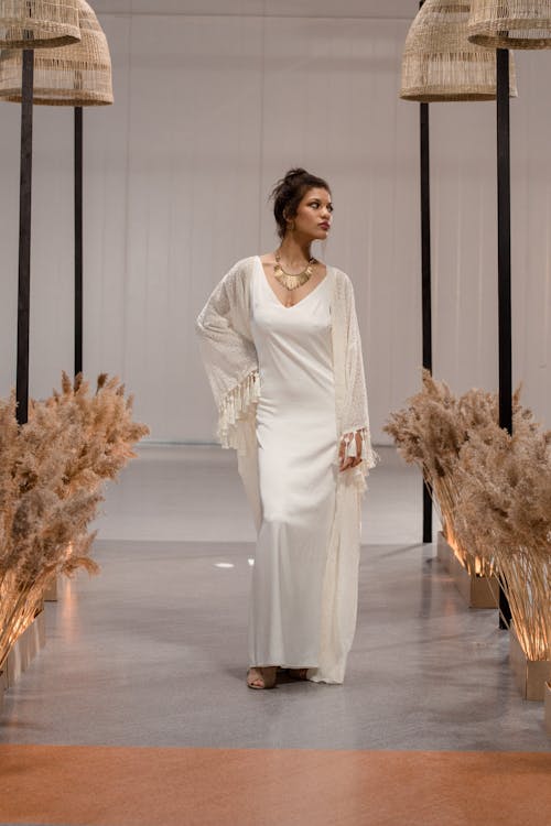 a fashion model showcases a stunning boho wedding dress. the dress is a combination of a silk ivory slip dress and a delicate lace kimono with tassels.  the model wears a simple gold necklace. the setting is minimalist and stylish, featuring pampas grass decor and neutral tones. the overall style evokes a sense of effortless elegance.
