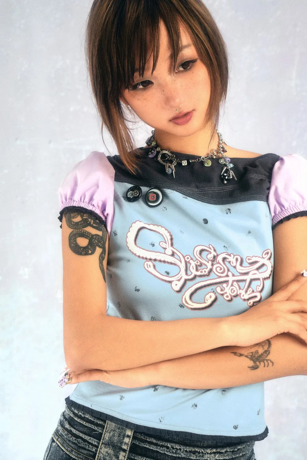 closeup portrait of a young asian woman with short brown hair and freckles. she's wearing a light blue and dark gray cropped top with purple sleeves and a graphic design. she also has a necklace and is wearing denim jeans. the girl has a snake tattoo on one arm and a scorpion tattoo on the other. she has her arms crossed and looks pensive, looking down. the background is a plain light gray.